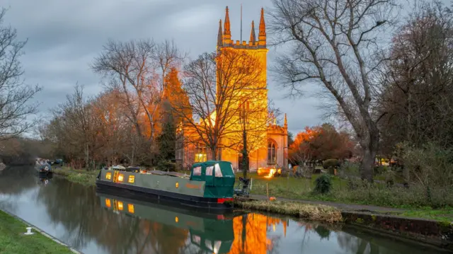Una iglesia con torre resplandece al atardecer, mientras el sol se refleja en la piedra. En primer plano, un canal con una barcaza verde. El agua está tranquila y las orillas están bordeadas de árboles.