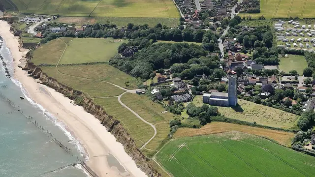 Una imagen aérea de una costa, una playa y un pueblo, que muestra campos directamente adyacentes a los acantilados y más allá de los campos una iglesia, un cementerio y viviendas.
