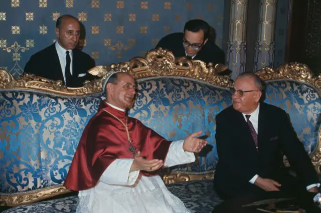 Papa 6. Paul ve Cevdet Sunay sohbet ediyor. Arkalarında takım elbiseli iki adam var.