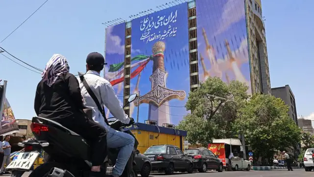 تہران میں موٹر سائیکل پر سوار دو افراد ایک عمارت پر بنے بڑے دیوارگیر نقش کے پاس سے گزر رہے ہیں جس میں خنجر اور داغے گئے میزائل دکھائے گئے ہیں