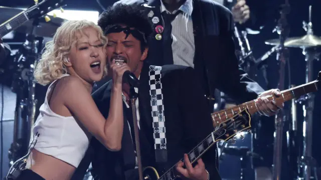 ギターを持ったマーズさんと、白いタンクトップ姿のロゼさんが、一つのマイクで歌っている。背後にはバックバンドが見える