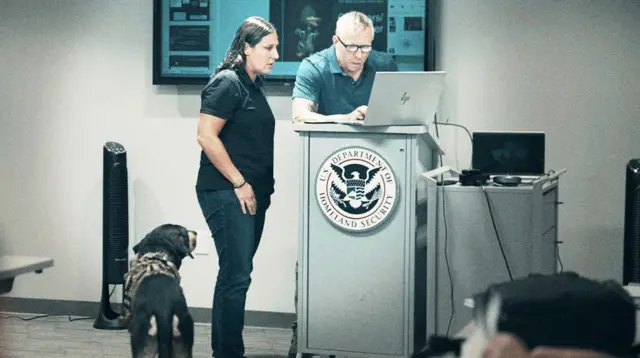 Женщина в черном поло и джинсах стоит рядом с Грегом Сквайром — мужчиной в синем поло. Сквайр в очках, за ноутбуком, стоит у трибуны с эмблемой DHS — Министерства внутренней безопасности США. Слева от них служебная собака.
