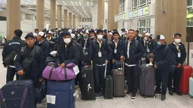 တောင်ကိုရီးယားကို ထွက်ခွါကြမယ့် မြန်မာလုပ်သားတွေ(ပုံဟောင်း)၊ အမျိုးသား အယောက် တစ်ရာထက်မနည်း နက်ပြာရောင် ဦးထုပ်၊ ဂျာကင်နဲ့ ဘောင်ဘီရှည်တွေ ဝတ်ဆင်ပြီး ကင်မရာဘက်ကိုကြည့်နေ၊ အချို့က မျက်နှာဖုံးနှာခေါင်းစည်း ဝတ်ထားပြီး လည်ပင်းမှာ တံဆိပ်ဆွဲထားသူအချို့နဲ့ ခရီးဆောင်အိတ်တွေကို ကိုင်ထားသူအချို့ကိုလည်း မြင်ရ၊ 