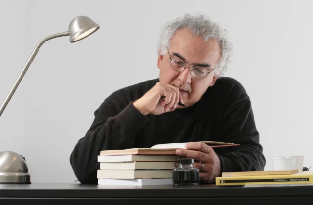 Milton Hatoum, um homem de cabelos grisalhos em um retrato em que aparece escrevendo em um caderno, sentado em uma mesa. Ele veste uma blusa preta e óculos.