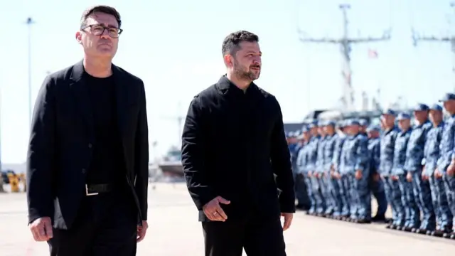 両首脳が海軍基地で並んで歩いている。二人とも黒い服を着ており、短い黒髪をしている。バーナム氏は眼鏡をかけている。背景には海軍関係者が並んでいる。
