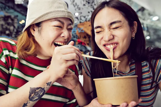 Woman feeding noodles with chopsticks to friend - stock photo Happy young woman feeding noodles with chopsticks to female friend at street