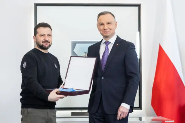 Зеленський і Дуда стоять у світлому офісі поруч із польським прапором; Зеленський тримає відкриту коробку з відзнакою. Обоє дивляться в камеру з посмішками.