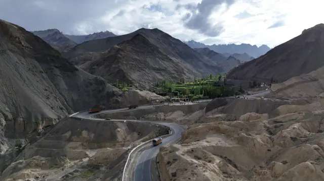 लद्दाख के स्कुरबुचन गांव की ओर जाने वाली एक पहाड़ी सड़क. इसमें घुमावदार सड़क दिखाई दे रही है, जिस पर कुछ ट्रक चल रहे हैं और चारों ओर बंजर पहाड़ हैं. बीच-बीच में हरे-भरे पेड़ों और वनस्पतियों का एक छोटा सा हिस्सा भी दिखाई दे रहा है. 