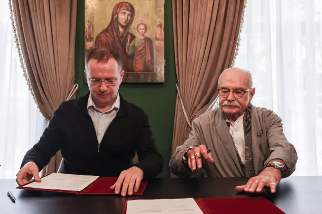 Двое мужчин — Владимир Мединский и Никита Михалков — сидят за столом в кабинете с зелеными стенами, на фоне висит икона Богородицы