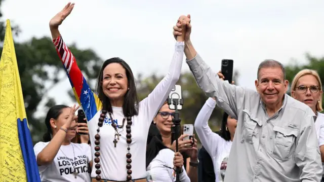 María Corina Machado viste una camiseta manga larga blanca. Sonríe mientras tiene los dos brazos en alto, dándole la mano izquierda a Edmundo González Urrutia, quien viste una camisa gris y también sonríe. Detrás de ellos se ven varias personas que parecen estar haciéndoles fotos o videos con teléfonos celulares en las manos.