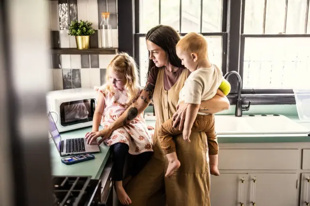 Mutfak tezgahına yaslanmış bir şekilde bilgisayarından iş halleden bir anne, aynı zamanda bir çocuğunu kucağında taşırken, tezgaha oturmuş diğer bir kız çocuğuyla da ilgileniyor. 