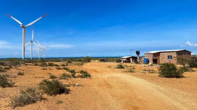 Terreno árido na Colômbia, com torres eólicas de um lado e construções do outro