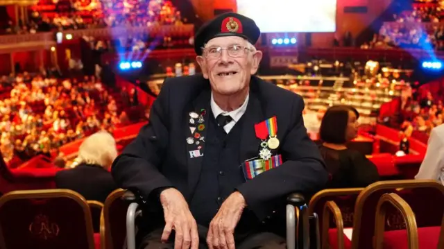 Jim Grant durante un concierto para celebrar el 80.º aniversario del Día de la Victoria en Europa. Está sentado en silla de ruedas y viste su uniforme de veterano. Detrás de él se ve el Royal Albert Hall, con público e iluminación.