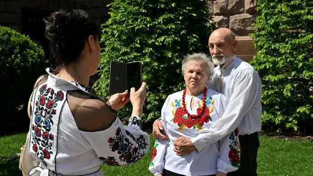 Молода жінка в яскравій вишитій сорочці фотографує на телефон двох літніх людей у вишиванках, які стоять, обійнявшись.