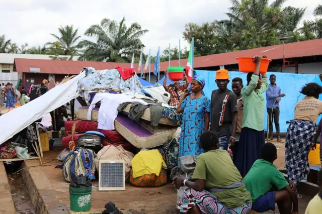 Impunzi ziri imbere y'inyubakwa ifise amabati atukura, imbere yaho hari amabendera, zimwe mu mpunzi zirahagaze, izindi ziricaye, abagore babiri bafise ama-bassins ku mutwe, bari iruhande y'umurundo w'amatungo yabo harimwo imatera, n'ibindi bitekeye mu mpuzu, hari n'impuzu zanikiye kuri iri matera, inyuma yabo habonena ihema risa n'ubururu