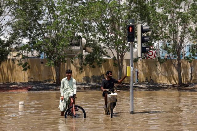 شخصان يركبان دراجتين، وسط مياه الفيضانات بعد هطول أمطار غزيرة في دبي - 19 أبريل/نيسان 2024