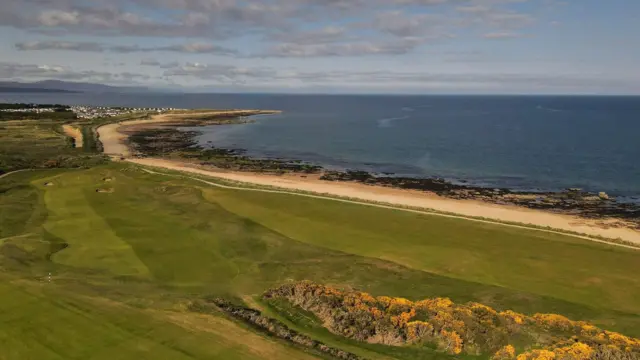 Una imagen aérea que muestra parte del campo de golf Royal Dornoch. Hay calles y búnkeres junto a una playa de arena dorada y un mar azul intenso.