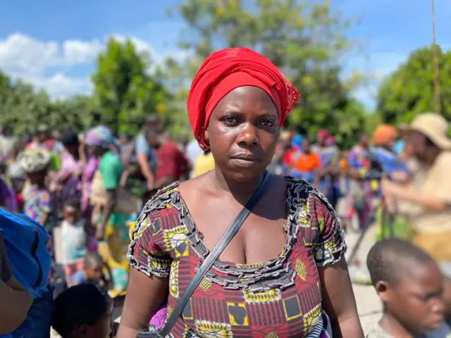 Umugore uteze igitambaro gitukura mu mutwe, inyuma ye hari umurongo w'abantu