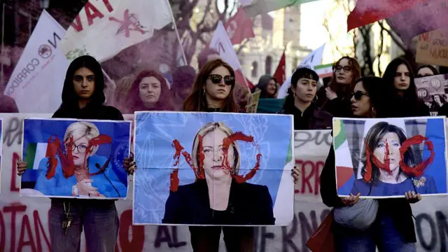 Ön tarafta üç kadın en önde Başbakan Meloni olmak üzere İtalyan politikacıların fotoğrafları üzerine kırmızı renkli hayır yazılı pankartlar tutuyor. Arkada altı kadın büyük bir pankart tutuyor. Arkalarında bayraklı bir kalabalık var. 