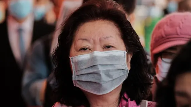 ਮਾਸਕ ਪਾਈ ਭੀੜ ਵਿੱਚ ਖੜ੍ਹੀ ਇੱਕ ਔਰਤ