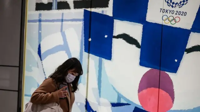 Женщина в маске на фоне плаката с эмблемой Токийской Олимпиады
