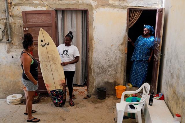 Khadjou Sambe est debout avec sa planche de surf alors qu'elle parle à son entraîneur Rhonda Harper sous le regard de sa mère