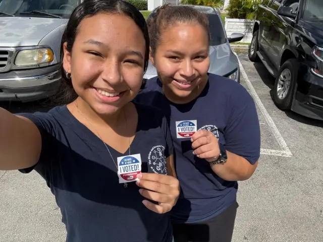 Christell and Leah take a selfie in their blue outfits, holding a sticker that says "I voted!"