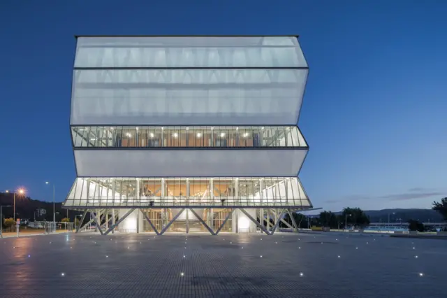 Edificio moderno iluminado al anochecer, con volúmenes superpuestos y fachada translúcida frente a una amplia plaza pavimentada.