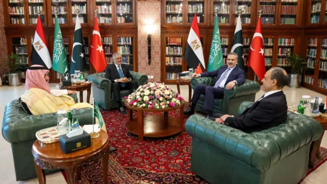 Четверо мужчин (главы МИД Пакистана, Египта, Саудовской Аравии и Турции) сидят в зеленых кожаных креслах. В комнате стоят флаги их стран, у стены — шкафы с книгами.