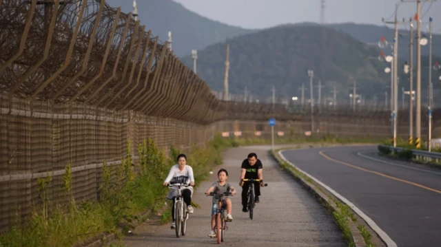 یک مرد و یک زن و یک کودک در کنار حصار طولانی در حال دوچرخه سواری هستند