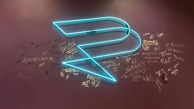 Una pared con una luz de neón con el logotipo de Production Park y autógrafos firmados de personas famosas que han estado en el espacio, incluidos Aitch, Editors y el elenco de Adolescence.