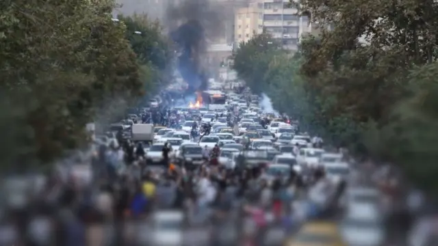 A wide street in Tehran blocked by vehicles, including cars and motorcycles, during the daytime on 21 September 2022. Dozens of people are gathered in front of and among the vehicles. A blur imposed by the BBC conceals the identities of people in the lower part of the image and nobody can be identified in the shot. A fire is burning in the background, with flames and dark black smoke billowing into the air. Green trees line both sides of the street. Tall buildings are in the background.
