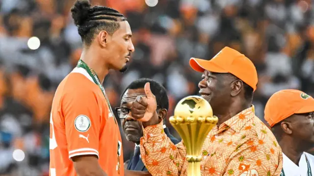 Le président ivoirien Alassane Ouattara (à droite), coiffé d'une casquette orange et vêtu d'un maillot à motifs orange, félicite le footballeur Sébastien Haller (à gauche), vêtu du maillot orange de l'équipe nationale, en levant le pouce après la victoire des Eléphants à la Coupe d'Afrique des Nations à Abidjan - 11 février 2024.