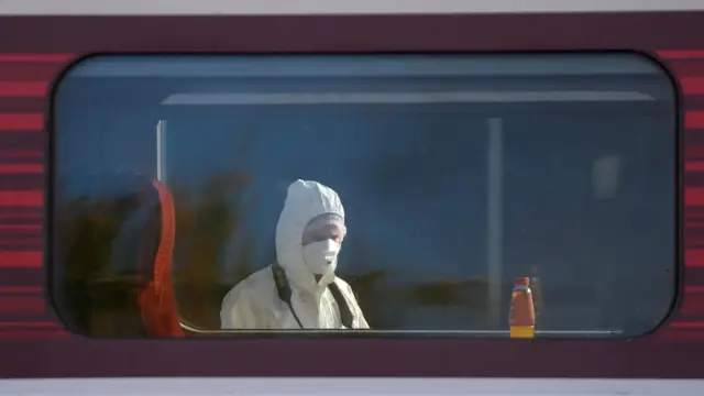 Cabeça e ombros de um perito policial são vistos através da janela de um vagão de trem. Ele usa capuz branco sobre a cabeça e máscara branca cobrindo nariz e boca.