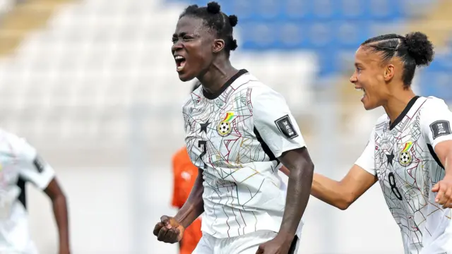 La princesse Marfo serre le poing droit et pousse un cri de joie en célébrant un but marqué pour le Ghana lors du match de barrage de la Coupe du monde féminine 2027 contre la Côte d’Ivoire. Derrière Marfo, on aperçoit sa coéquipière ghanéenne Chantelle Boye-Hlorkah qui se joint à la fête. On voit les deux joueuses de la taille aux épaules, vêtues de maillots blancs du Ghana ornés sur le devant d’un motif élaboré représentant une toile d’araignée multicolore. À l'arrière-plan, floue, on aperçoit une tribune vide dont les sièges sont bleus et blancs.