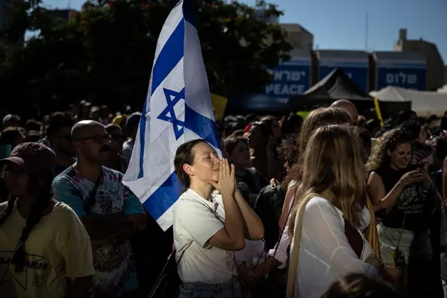 Bir kadın ellerini dua eder gibi kapamış, gözleri de kapalı, etrafından çok sayıda kişi var.