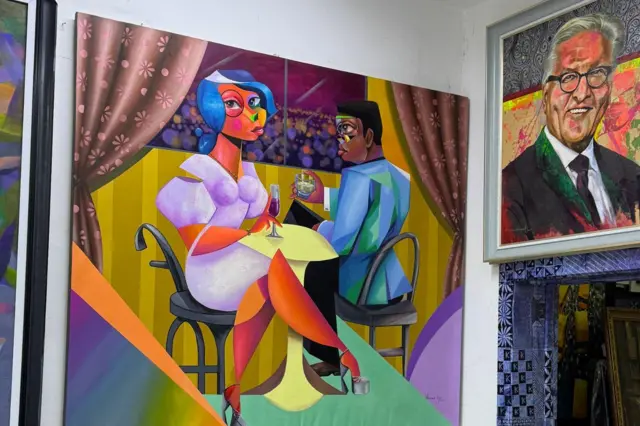 A large colourful painting hangs on a white gallery wall, depicting two stylised figures seated at a small table with drinks, framed by patterned curtains and geometric blocks of yellow, purple, blue and green. To the right, a framed portrait painting of a person wearing glasses is displayed above additional artworks, with more paintings partially visible at the edges of the image.
