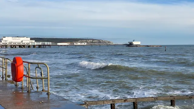 Un paisaje marino con varias olas rompiendo contra el malecón. A lo lejos se ve un muelle con dos edificios blancos y un acantilado. El cielo es azul y brilla el sol.