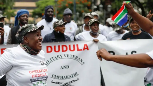 A woman in a white Operation Dudula T-shirt and wearing camouflage cap and scarf smiles as she participates in a demonstration by the anti-migrant group. Others are similarly dressed and a South African flag can be seen.