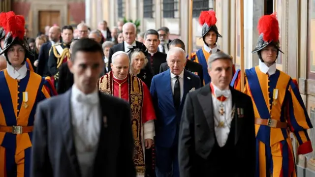 赤と白の法衣の教皇と紺のスーツ姿の国王が並んで歩き、その後ろに黒衣のカミラ英王妃が続く。左右に、オレンジと紺の縦じまの伝統的な服を着たスイス衛兵が並んでいる