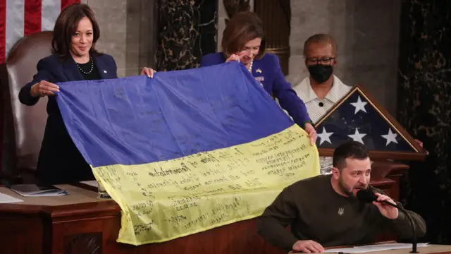 Kamala Harris e a presidente da Câmara, Nancy Pelosi seguram uma bandeira ucraniana enquanto o presidente ucraniano Volodymyr Zelensky faz um discurso em uma reunião conjunta do Congresso dos EUA em dezembro de 2022