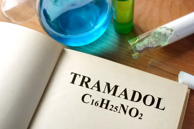 Sebuah buku terbuka dengan halaman bertuliskan tramadol dan kode kimia di atasnya berderet cairan berwarna biru.