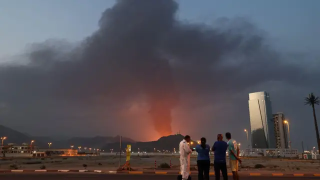 صورة من الأعلى - عمال أجانب ينظرون إلى عمود كثيف من الدخان الأسود يتصاعد عقب انفجار في المنطقة الصناعية بالفجيرة في 3 مارس/آذار 2026. أجبرت الغارات الإيرانية على دول الخليج المجاورة منذ 28 فبراير/شباط، عقب الهجوم الأمريكي الإسرائيلي، الإمارات العربية المتحدة على إغلاق مجالها الجوي، مما فاجأ المسافرين الذين ظنوا أنهم متجهون إلى واحدة من أكثر الوجهات السياحية أمانًا في المنطقة.