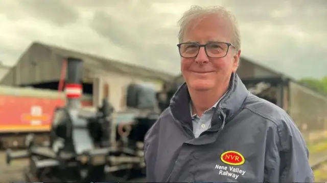 Michael Purcell de pie frente a una locomotora y unas cocheras. Lleva un impermeable gris con la marca roja y amarilla del ferrocarril a la izquierda. Sonríe a la cámara. Lleva gafas de montura cuadrada y cabello corto y blanco.