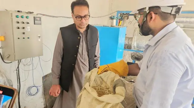 ڈاکٹر جبران خالق پاکستان کے شہر صالح پٹ میں منعقدہ ایک نمائشی تقریب میں۔
لیبارٹری کے ماحول میں سفید قمیص اور ذاتی حفاظتی ساز و سامان (PPE) پہنے ایک شخص سفید تھیلا تھامے ہوئے ہے، جبکہ ڈاکٹر خالق اس کے مندرجات کا جائزہ لے رہے ہیں۔
