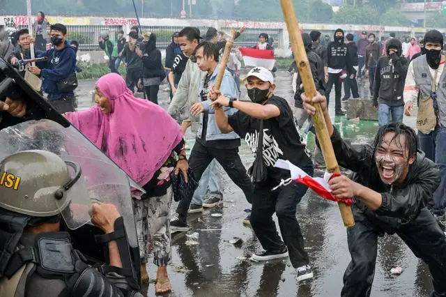 Beberapa orang pria mengangkat bambu yang terikat bendera merah putih sedang berhadapan dengan kelompok polisi.
