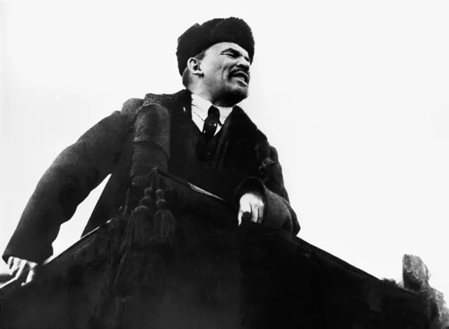 198 tarihli fotoğrafta Lenin, bir kürsüden konuşma yaparken görülüyor. Başında kalpak bulunuyor. 