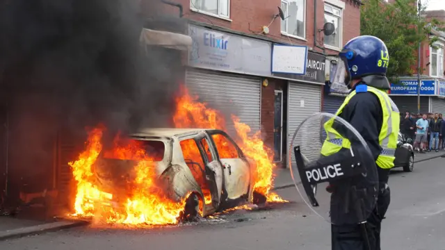 A police officer carrying a riot shield and wearing a helmet looks on as flames rise from a car on Middlesbrough's Parliament Road. A small crowd on people can be seen on a nearby corner.