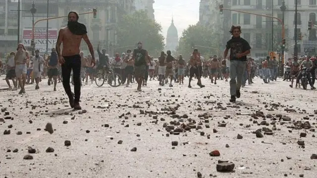 Pedras e destroços na rua, com manifestantes ao fundo