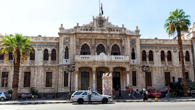 ساختمان ایستگاه راه‌آهن حجاز در دمشق. در مقابل بنای تاریخی خودروها و در دو سوی آن درختان نخل دیده می‌شود.
